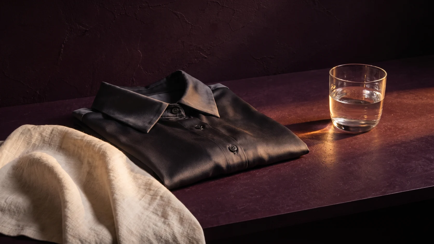 Stilleben med en vikt mörk skjorta, en handduk och ett glas vatten i varmt ljus.